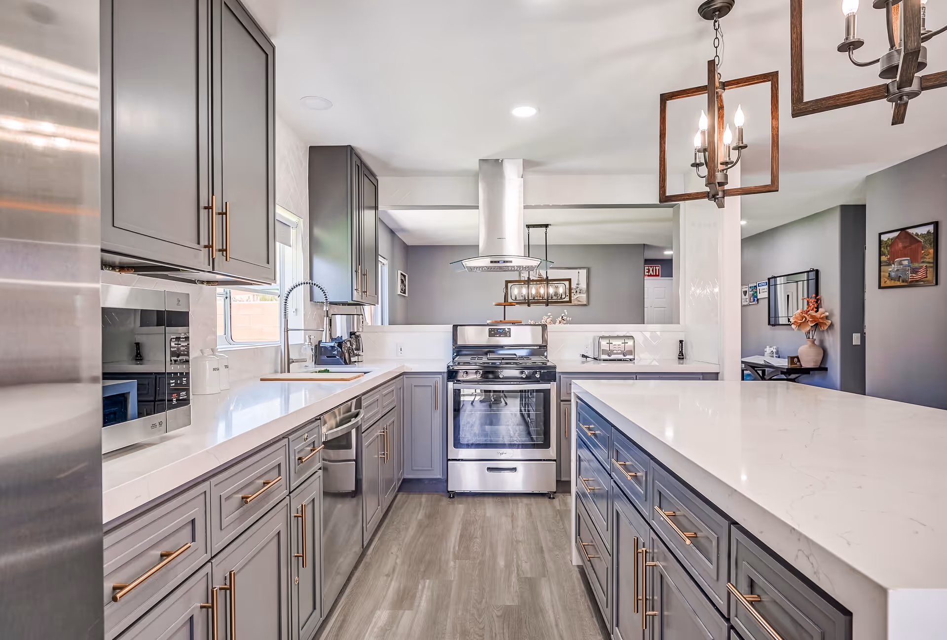 Modern kitchen with gray cabinets, white countertops, stainless steel appliances including a refrigerator, microwave, dishwasher, and stove with a range hood. The kitchen features a large island with drawers and pendant lighting above. The floor is light wood, and there is a window above the sink letting in natural light. The kitchen opens into a living area with gray walls and decorative elements.
