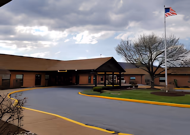 Exterior view of a single-story building with a brown roof and brick walls, featuring a covered entrance driveway. There is a flagpole with an American flag and another flag, along with a tree and some landscaping near the entrance.