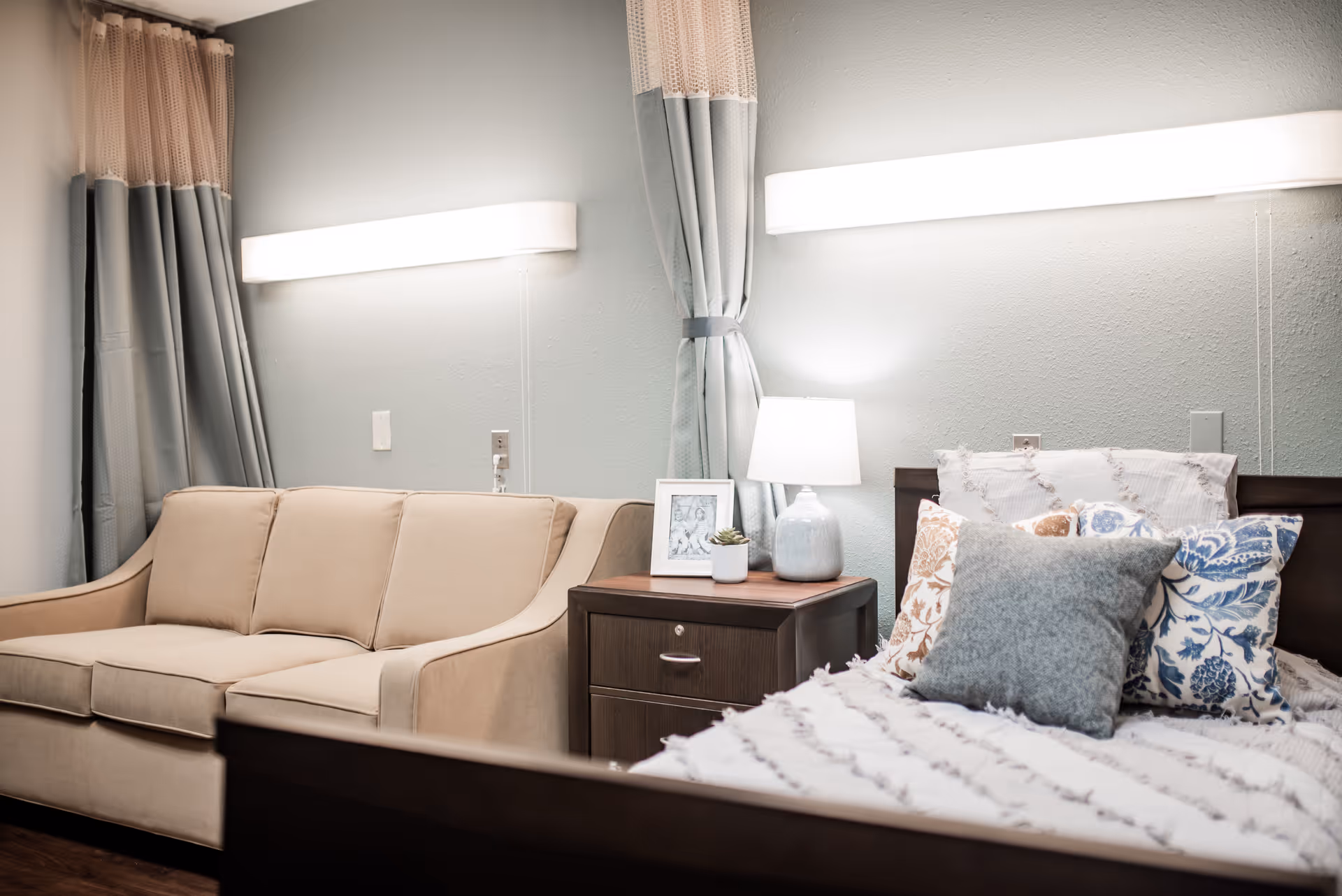 A cozy nursing facility room featuring a beige sofa, a wooden nightstand with a lamp, a small framed picture, and a potted plant. The bed is neatly made with multiple decorative pillows in various patterns and colors. The walls are light gray with two horizontal wall lights above the sofa and bed, and curtains hang in the corner.