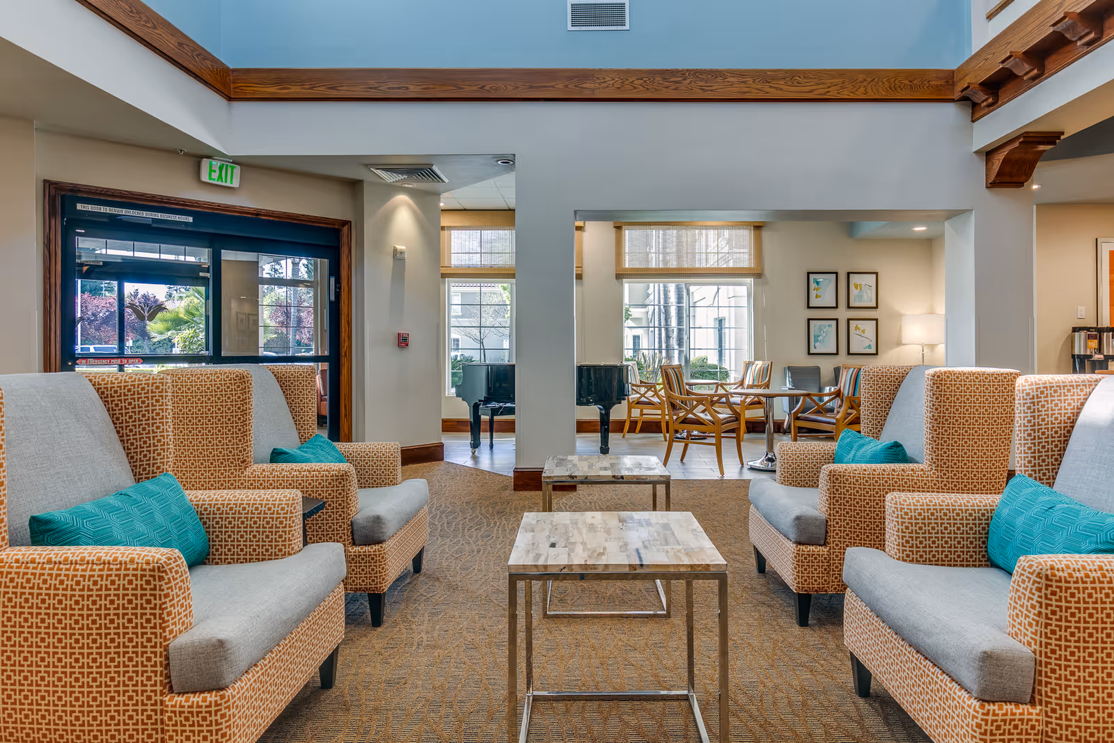 A bright and spacious senior living facility lounge area with patterned armchairs featuring teal cushions arranged around two small marble-top tables. Large windows let in natural light, and a black grand piano is visible in the background near a seating area with wooden chairs and a table. The room has high ceilings with wooden beams and neutral-colored walls.