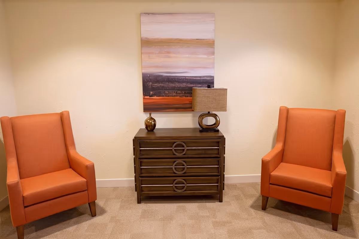 A cozy seating area with two orange upholstered armchairs facing forward, separated by a dark wooden chest of drawers. On top of the chest are a modern table lamp with a beige shade and a small decorative vase. Above the chest hangs a large abstract painting with horizontal bands of muted colors including beige, brown, and orange. The walls are light beige and the floor is carpeted in a neutral tone.