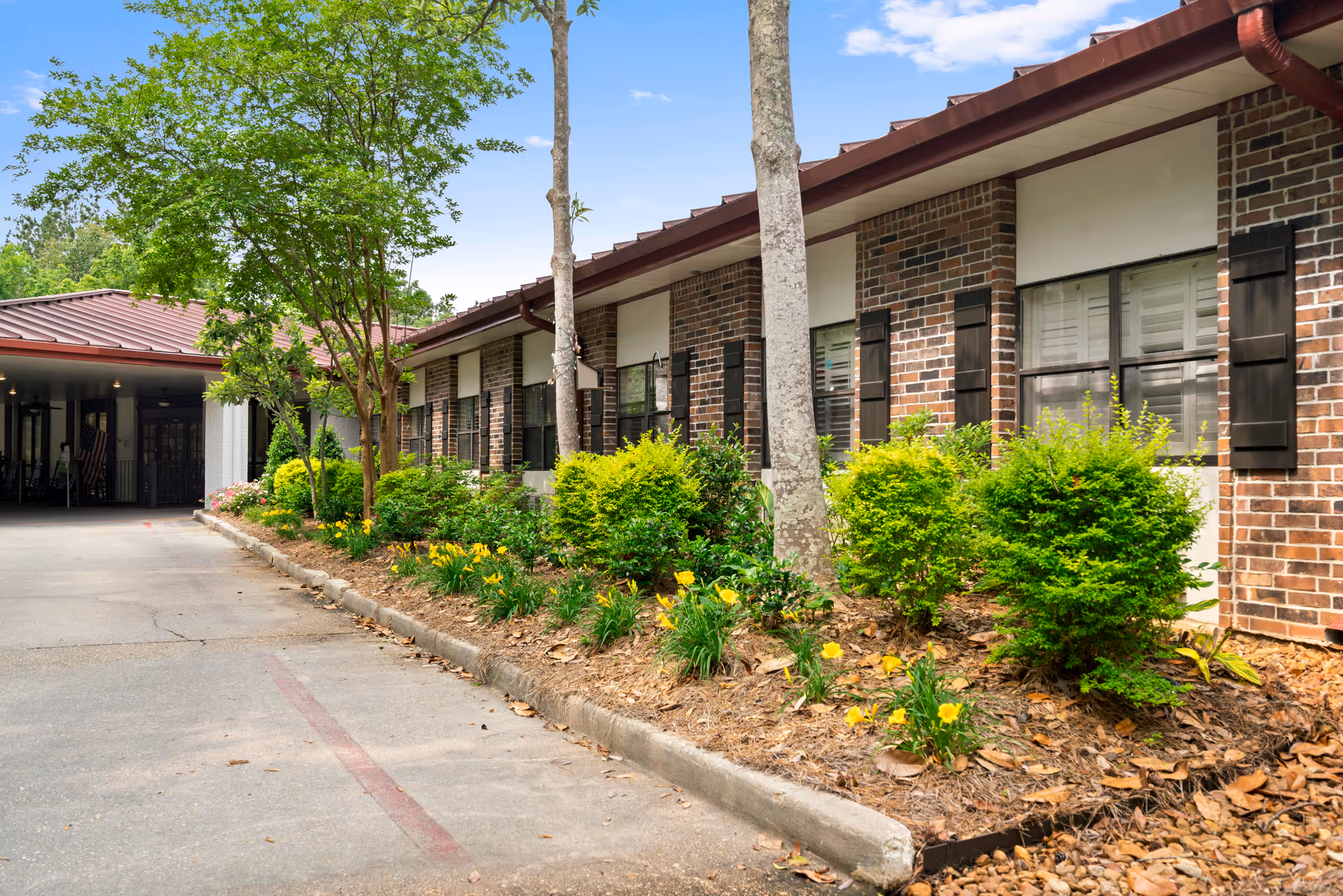 Exterior view of a single-story brick building with multiple windows, black shutters, and a red roof. There is a landscaped garden bed with green shrubs, yellow flowers, and trees along the building. A paved driveway leads to a covered entrance area.