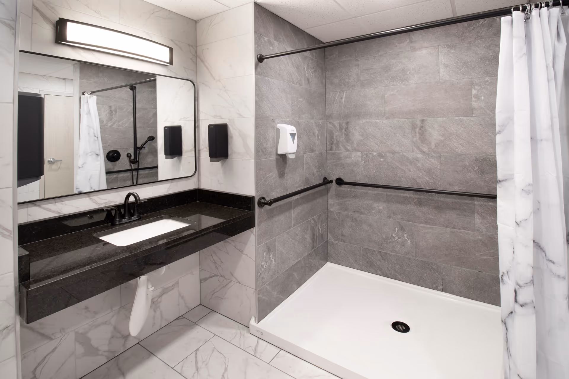A modern bathroom featuring a large rectangular mirror above a black countertop with a white sink. The walls are tiled with white marble-patterned tiles, and the shower area has gray stone tiles with black grab bars and a white shower curtain.