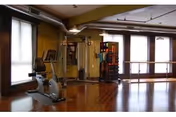 A fitness room with wooden floors and large windows letting in natural light. The room contains exercise equipment including a stationary bike and a multi-functional weight machine. There is also a ballet barre along one wall.