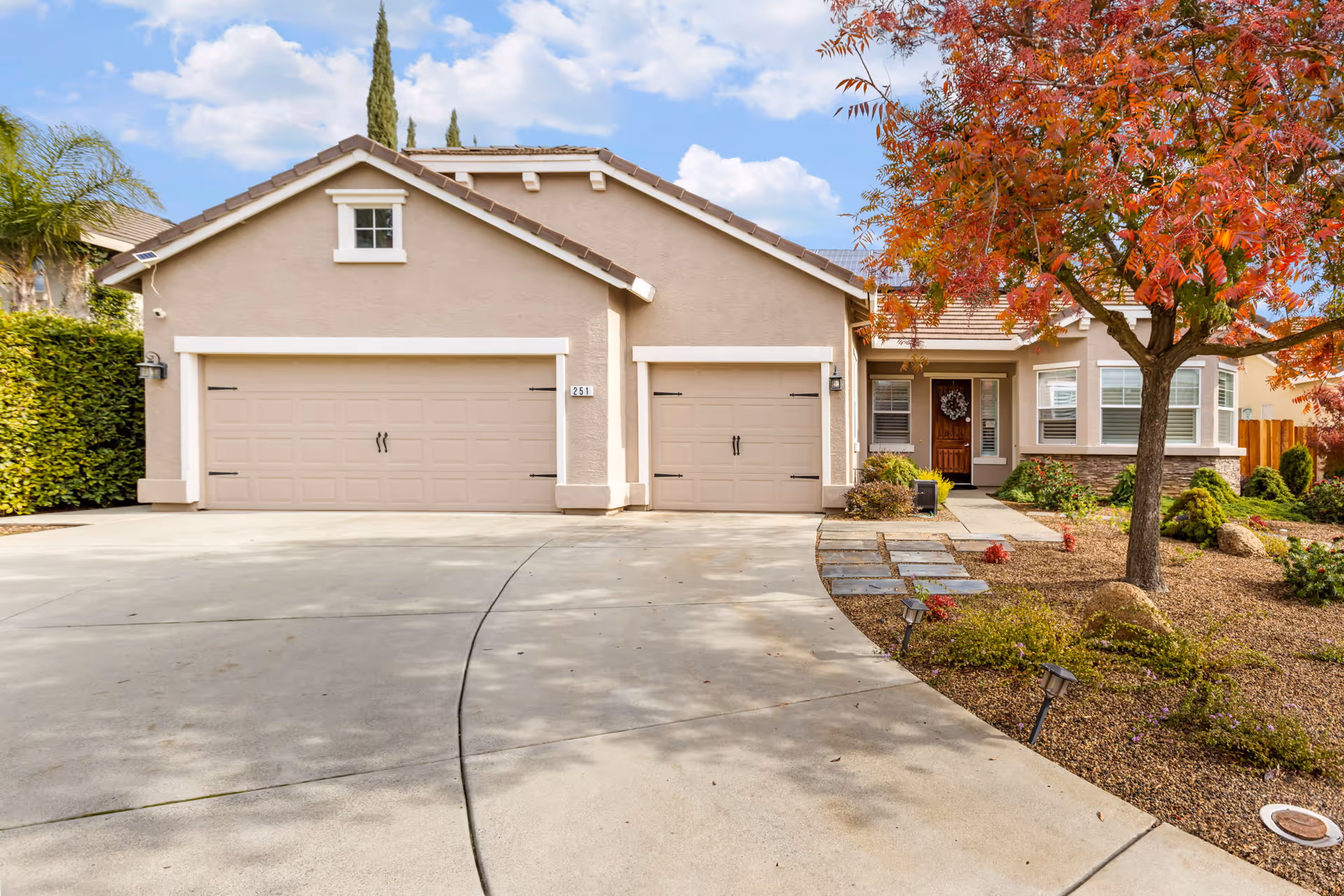 Front exterior of a single-story house with a three-car garage, curved driveway, walkway, and a red-leaved tree.