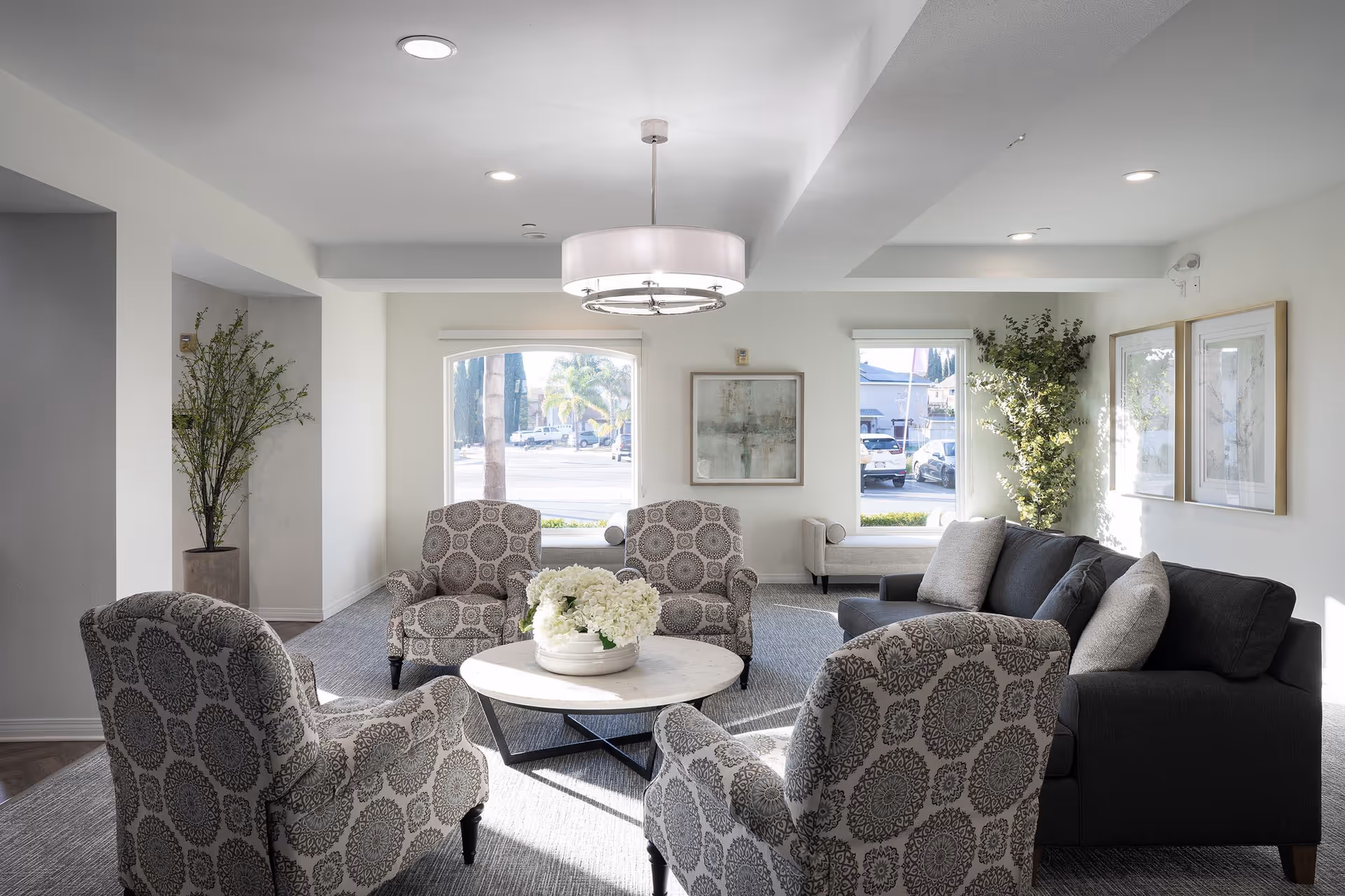 A bright and modern living room area with four patterned armchairs arranged around a round white coffee table with a flower arrangement. There is a dark gray sofa with cushions to the right, two large windows letting in natural light, framed artwork on the walls, and tall green plants in the corners.