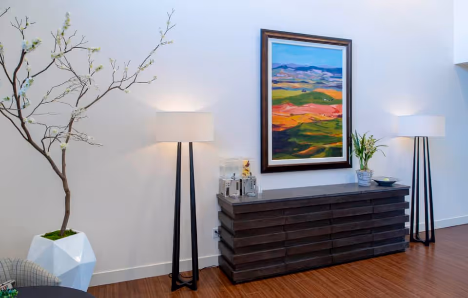 A modern interior space featuring a dark wooden sideboard with decorative items including a small plant and a glass container. Above the sideboard hangs a colorful landscape painting. On either side of the sideboard are two tall floor lamps with white shades. To the left, there is a large white geometric planter holding a branch with white blossoms. The floor is wooden and the walls are white.
