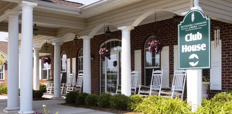 Exterior view of a brick building with white columns and a covered porch featuring white rocking chairs and hanging flower baskets. A green sign near the entrance reads 'Belvedere Commons of Seneca Club House' with an arrow pointing to the right.