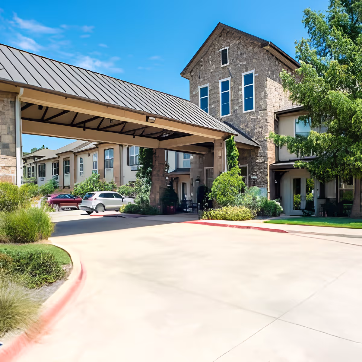 Exterior view of Discovery Village At Southlake showing a stone building with multiple windows, a covered driveway entrance, surrounding greenery, and parked cars under a clear blue sky.
