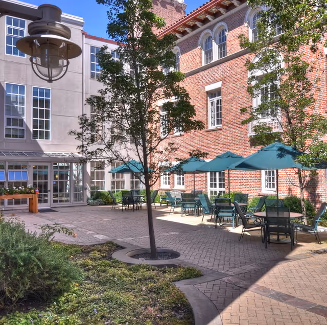 Sunlit courtyard with patio tables and green umbrellas between a red brick building and a modern building.