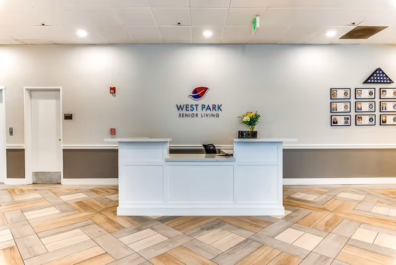 Reception desk area at West Park Senior Living with a white counter, a chair, a vase with yellow flowers, and a wall displaying the facility's logo. The floor has a patterned wood and tile design, and there are framed photos and a folded American flag on the wall to the right.