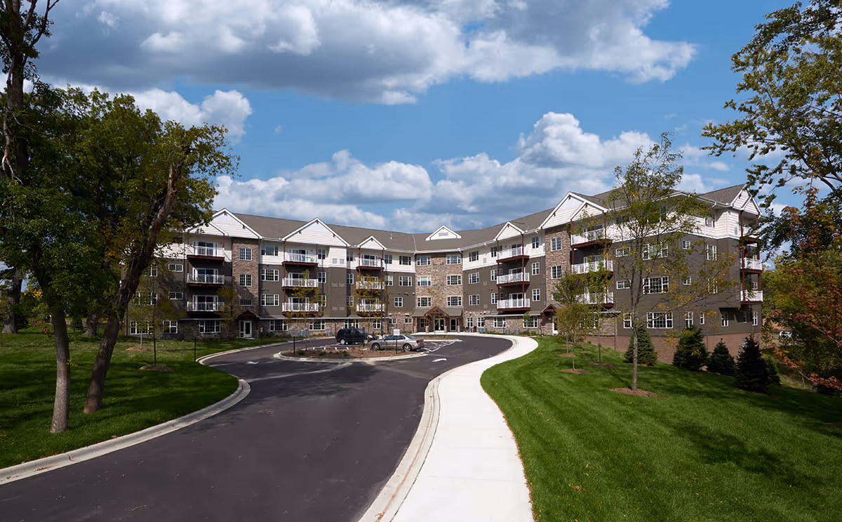 Exterior view of a multi-story senior living facility building with a curved driveway, green lawns, and trees under a partly cloudy blue sky.