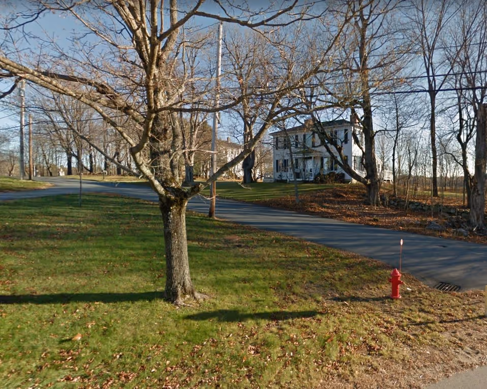 A rural scene showing a two-story white house with black shutters situated on a grassy area with leafless trees and a paved road curving around the property. A red fire hydrant is visible near the road.