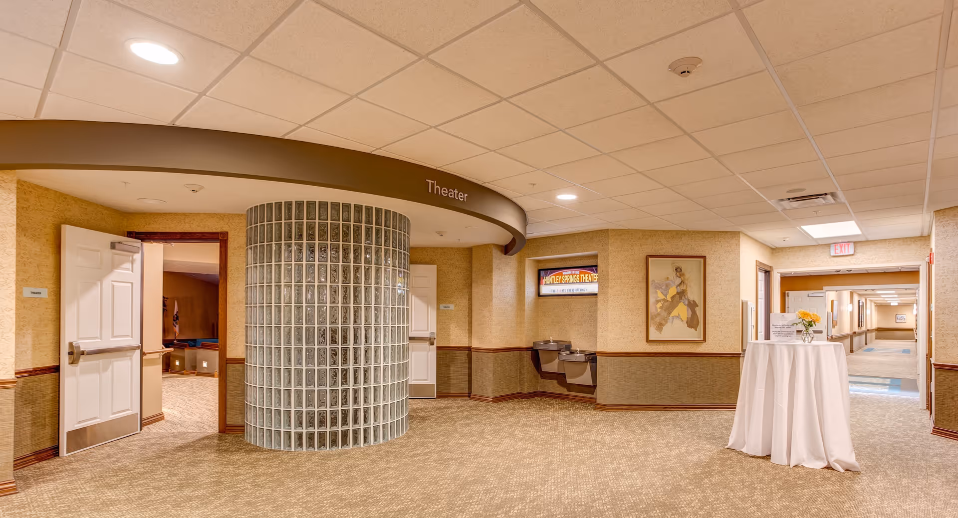 Interior hallway of Huntley Springs Retirement Resort featuring a curved glass block wall with an entrance to a theater room. The hallway has beige walls with a chair rail, carpeted floor, ceiling tiles with recessed lighting, two water fountains, a framed artwork, and a small round table covered with a white cloth and a flower vase. There is an exit sign above a door at the end of the hallway.
