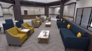 A modern senior living facility common area with multiple seating arrangements including blue sofas with yellow pillows and gray armchairs with yellow cushions. The room features large windows, a patterned carpet, and a neutral color palette with dark wood accents.