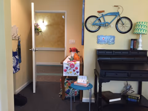 Interior view of a hallway in a senior living facility with an open door leading to another room. On the right side, there is a black wooden desk with books and decorative items on and under it. Above the desk, a blue bicycle is mounted on the wall along with a decorative sign and a green patterned lamp. A small round blue table with books is placed next to the desk. The walls are painted light yellow and the floor is carpeted.