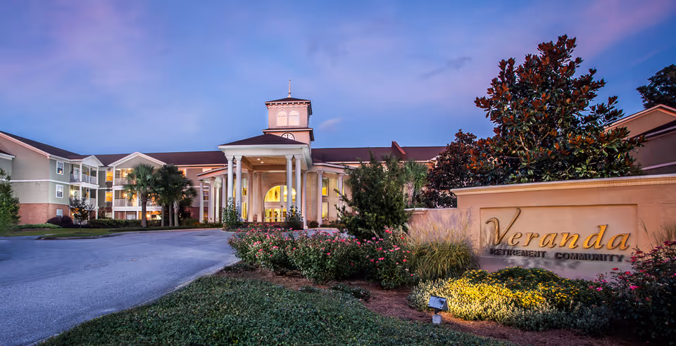 Front entrance of the Veranda of Pensacola retirement community with a landscaped driveway, illuminated portico, and signage at dusk.