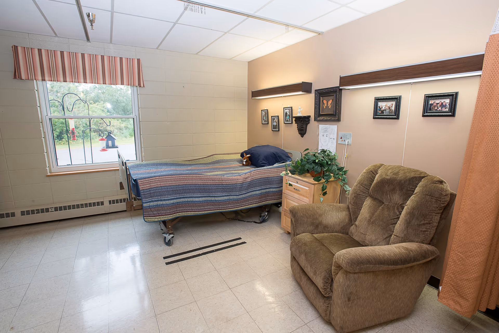 A small, cozy room with a single hospital-style bed covered with a striped blanket, a brown cushioned armchair, a wooden nightstand with a green potted plant, and several framed pictures on the wall. There is a window with a striped valance letting in natural light and a curtain partially visible on the right side.