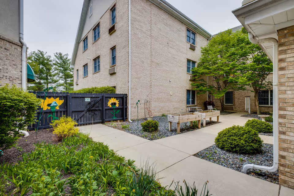 Outdoor courtyard area at West Lafayette Assisted Living featuring a paved walkway, green shrubs, trees, and raised garden beds. The courtyard is surrounded by a beige brick building and a black wooden fence with painted sunflowers and a small exit sign.