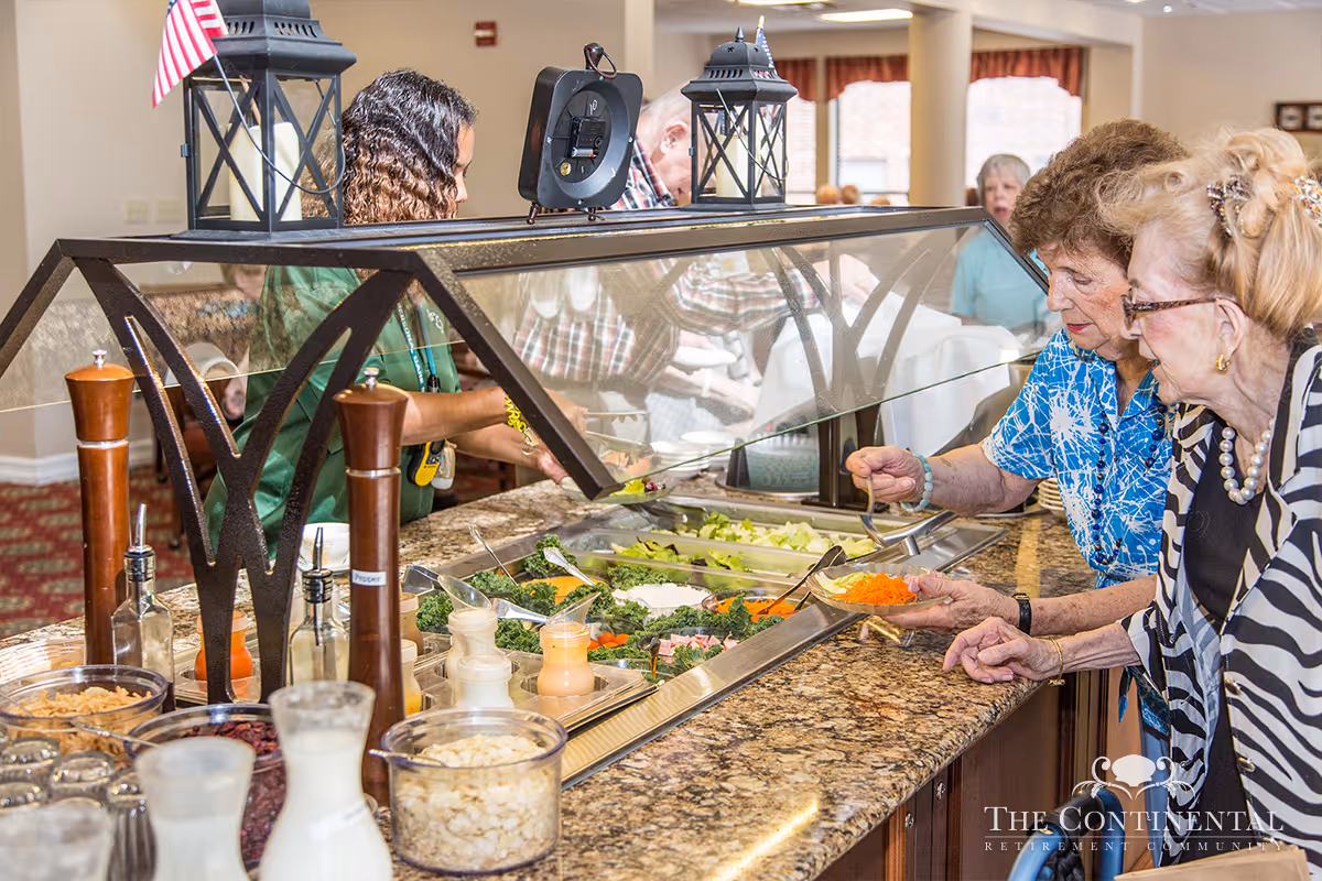 Two elderly women serving themselves salad from a buffet salad bar in a dining area, with a staff member assisting behind the counter. Various salad ingredients and dressings are visible on the counter.