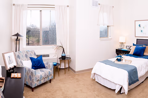 A bright and cozy bedroom featuring a single bed with white and blue bedding, a bedside table with a lamp and decorative items, a patterned armchair with a blue pillow, a small round side table with a plant, and a large window with white curtains letting in natural light.