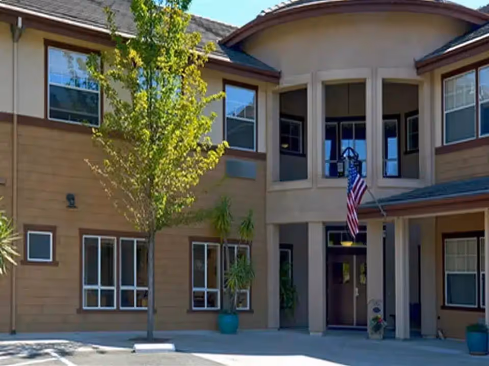 Entrance of a two-story assisted living building with windows, an American flag, and a small tree in front.