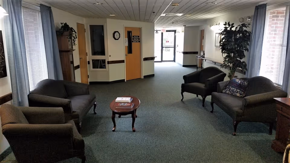 A quiet seating area in a senior living facility hallway with two armchairs and two loveseats arranged around a small wooden coffee table. The space has green carpet, light-colored walls, large windows with blue curtains, a potted plant, and doors leading to other rooms. A clock and signage are visible on the walls.
