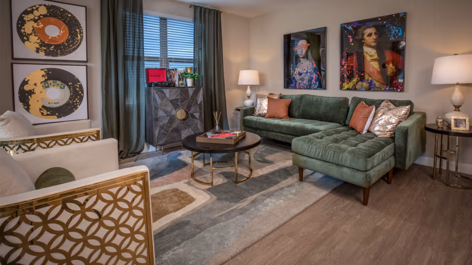Contemporary living room with a green sectional sofa, gold-accent chairs, round coffee table, patterned rug and framed artwork on the walls.