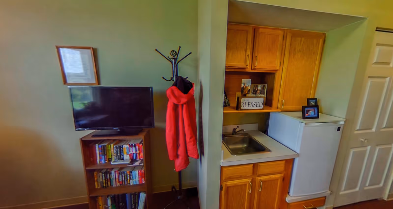 Small apartment interior showing a kitchenette with a sink and mini-fridge next to wooden cabinets, a coat rack with a red jacket, and a TV on a bookshelf.