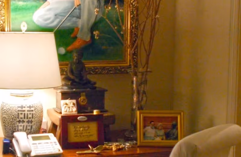 A cozy interior corner featuring a table with a decorative lamp, a telephone, a small statue of a seated figure, framed photographs, and a painting of a person playing golf on the wall behind.