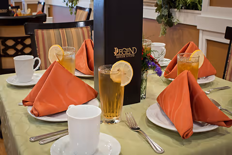 A dining table set for four with orange folded napkins on white plates, glasses of iced tea with lemon slices, white coffee cups on saucers, silverware, and a black menu with the text 'Legend Senior Living'. The table has a light green tablecloth and there are chairs with striped upholstery around it.