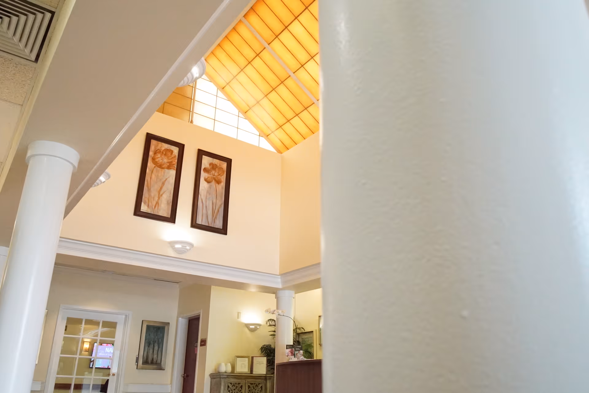 Interior view of a senior living facility lobby with a high ceiling and a large skylight. Two framed floral artworks hang on the wall above a reception desk. White columns and soft lighting create a welcoming atmosphere.