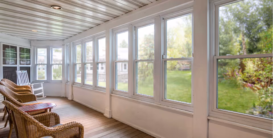 Bright enclosed porch with wicker chairs and a rocking chair facing a row of windows overlooking a green yard.