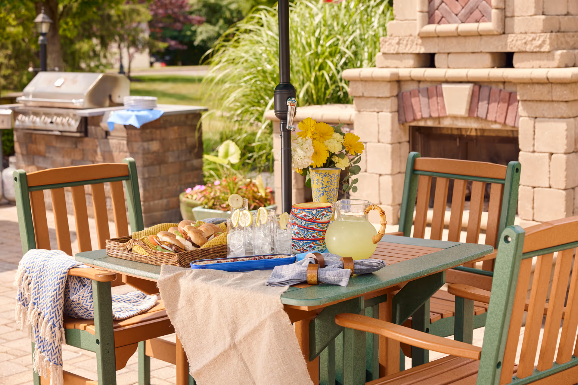 Outdoor patio dining area with a wooden table and four chairs. The table is set with a pitcher of lemonade, glasses with lemon slices, a basket of sandwiches, colorful bowls, and a vase with yellow and white flowers. In the background, there is a built-in stone grill and a stone fireplace surrounded by greenery.