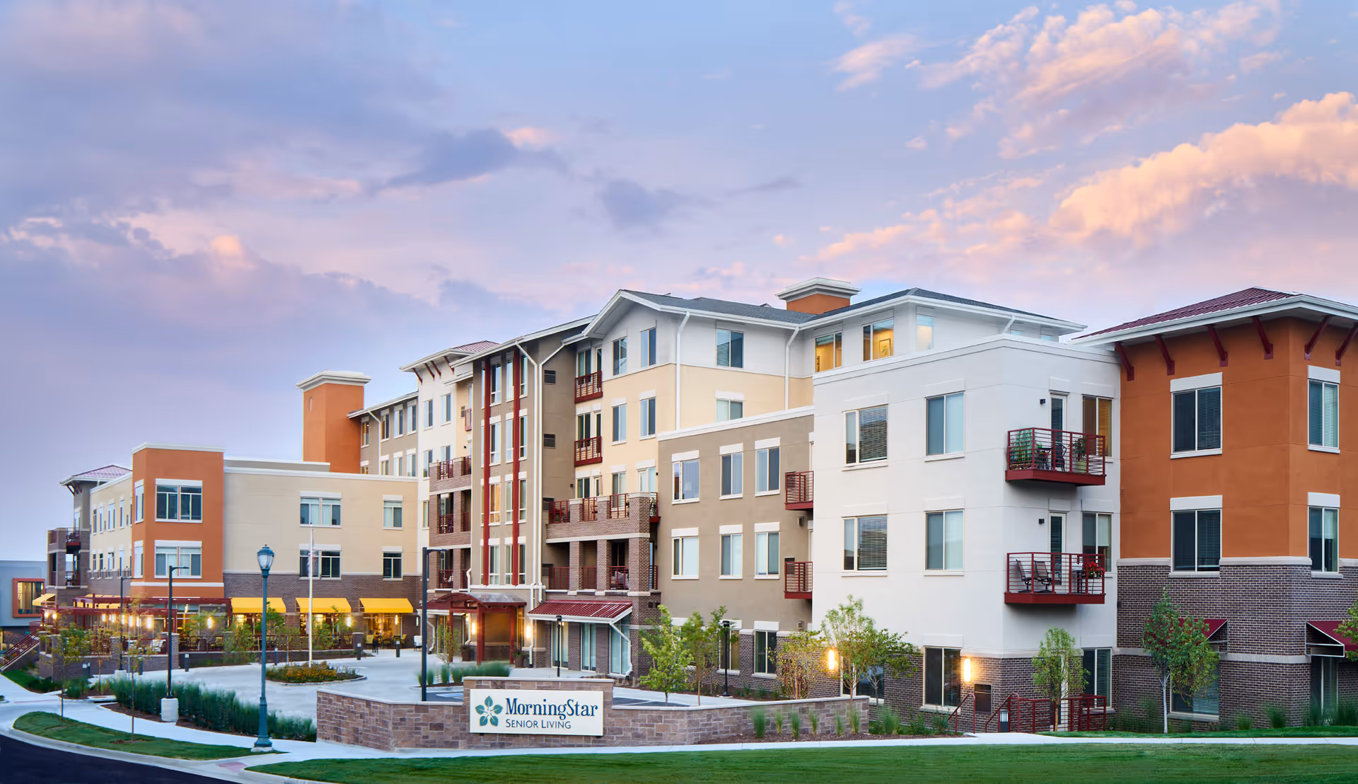 Exterior view of a modern multi-story senior living facility building with a mix of beige, white, and orange tones, featuring balconies, large windows, landscaped greenery, and a sign in front that reads 'MorningStar Senior Living'. The sky is partly cloudy with a soft pink and blue hue during sunset or sunrise.