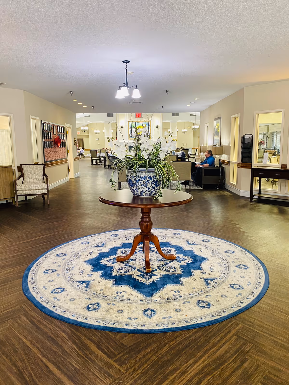 A senior living facility lobby with a round table holding a blue-and-white vase of flowers on a circular rug and seating areas in the background.