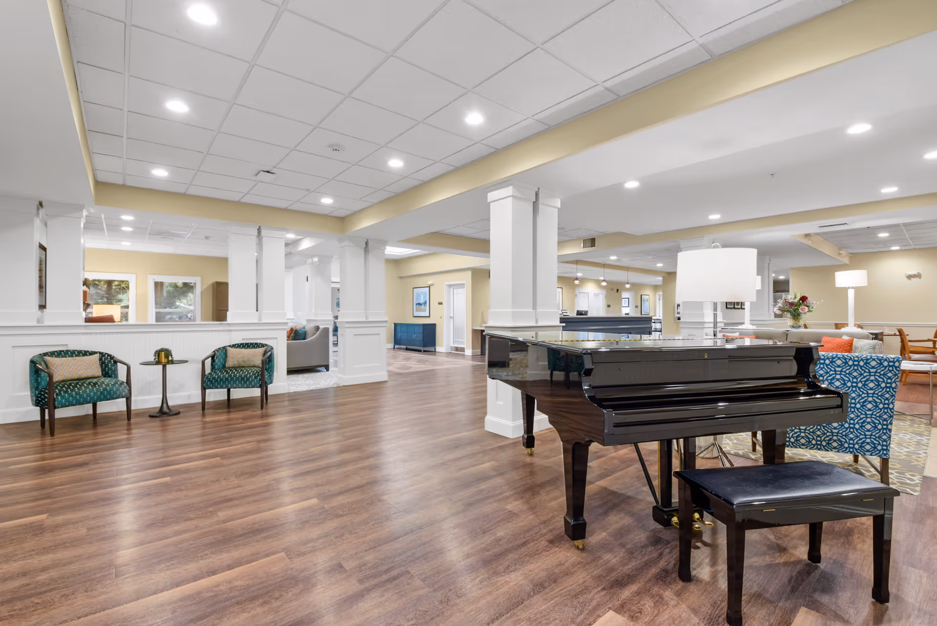 A spacious and well-lit common area in a senior living facility featuring a black grand piano with a matching bench, two green patterned armchairs with a small round table between them, and additional seating areas with armchairs and lamps in the background. The room has wood flooring, white pillars, and a ceiling with recessed lighting.