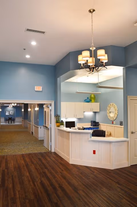 Interior view of a reception area in a senior living facility with a white reception desk, computer, and chairs. The walls are painted blue with white cabinetry and a decorative round mirror. A chandelier with five lights hangs from the ceiling. In the background, a hallway with carpeted flooring and additional seating is visible.