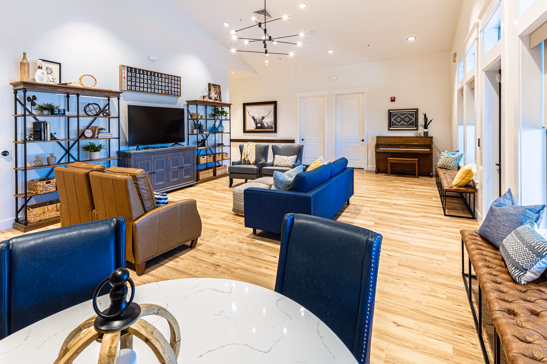 Spacious, brightly lit communal living room with blue sofas, leather armchairs, a TV on a console, shelving, and a piano.