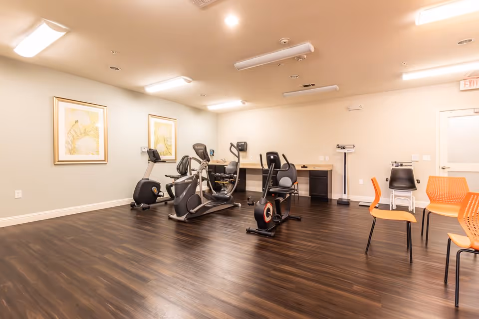 A fitness room in an assisted living facility with exercise equipment including stationary bikes and an elliptical machine. The room has wood flooring, light-colored walls with two framed pictures, and several orange chairs arranged near the right side. There is also a scale and a desk area along the back wall.