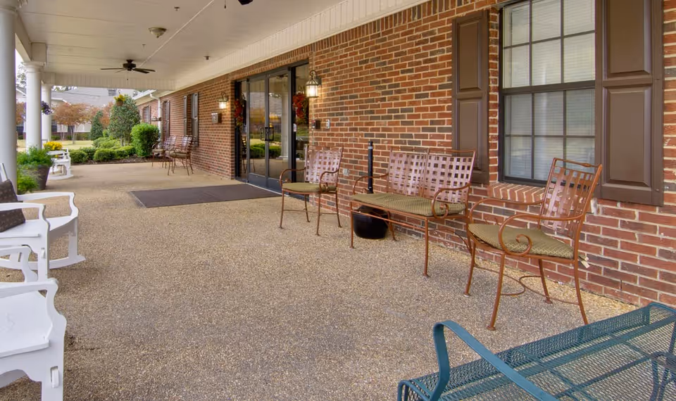 Covered outdoor patio area with several metal and wooden chairs and benches arranged along a brick wall with windows and a glass door entrance. The patio overlooks a landscaped garden with shrubs and trees.