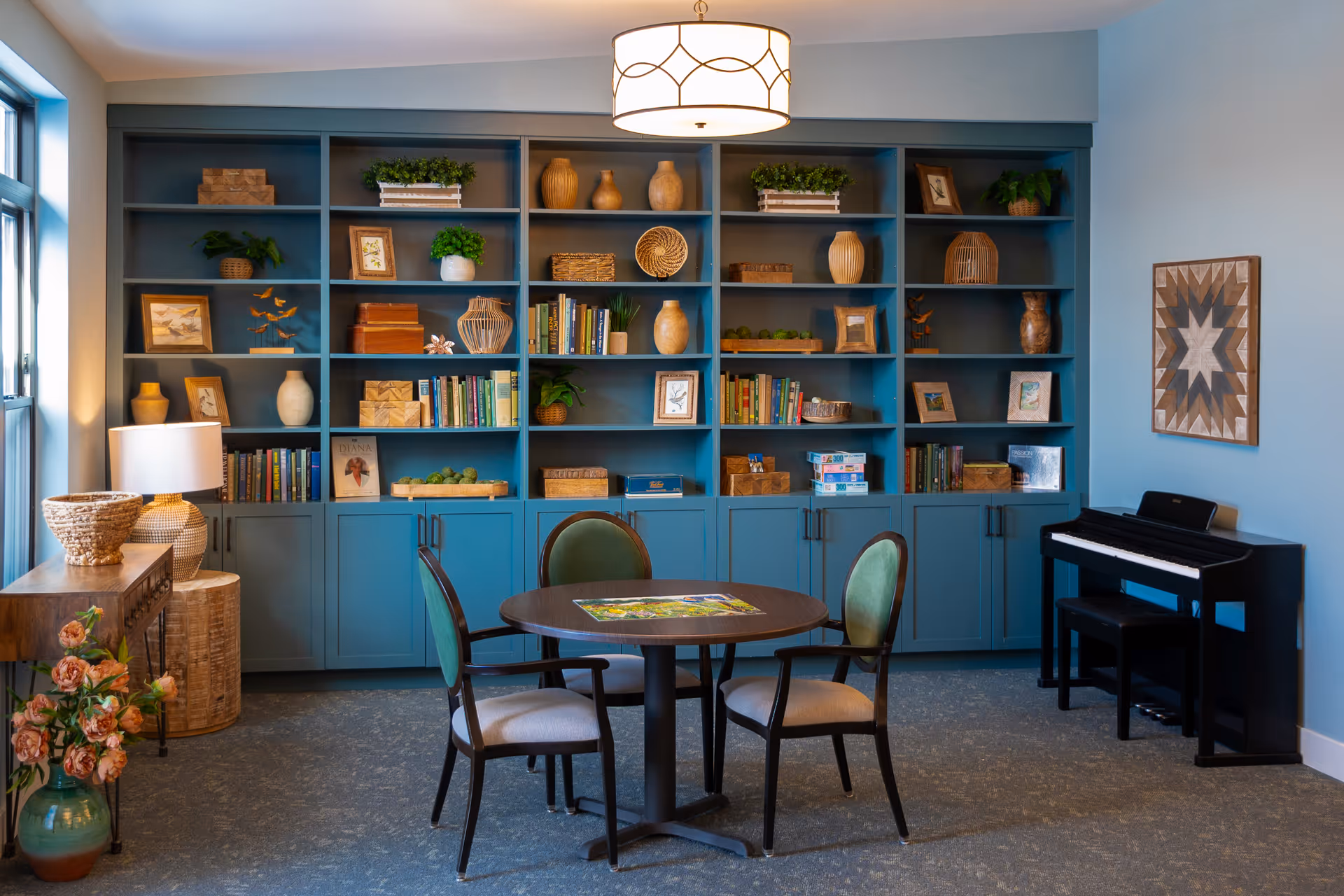 A well-appointed senior living common room with teal built-in bookshelves, a round table with four chairs, and a digital piano.