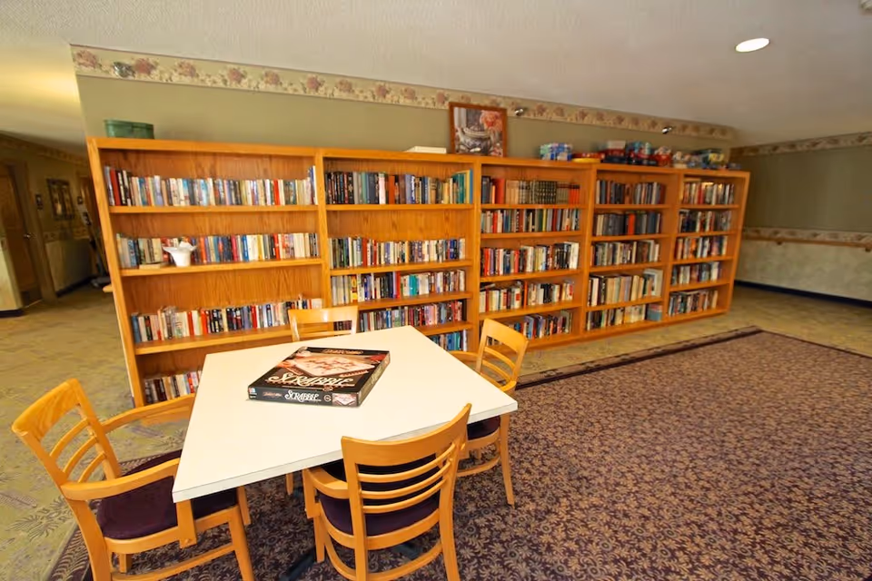 A cozy reading and game area in a senior living facility with a white square table surrounded by four wooden chairs with purple cushions. On the table is a Scrabble board game. Behind the table are large wooden bookshelves filled with books. The room has a patterned carpet and light green walls with floral wallpaper border near the ceiling.