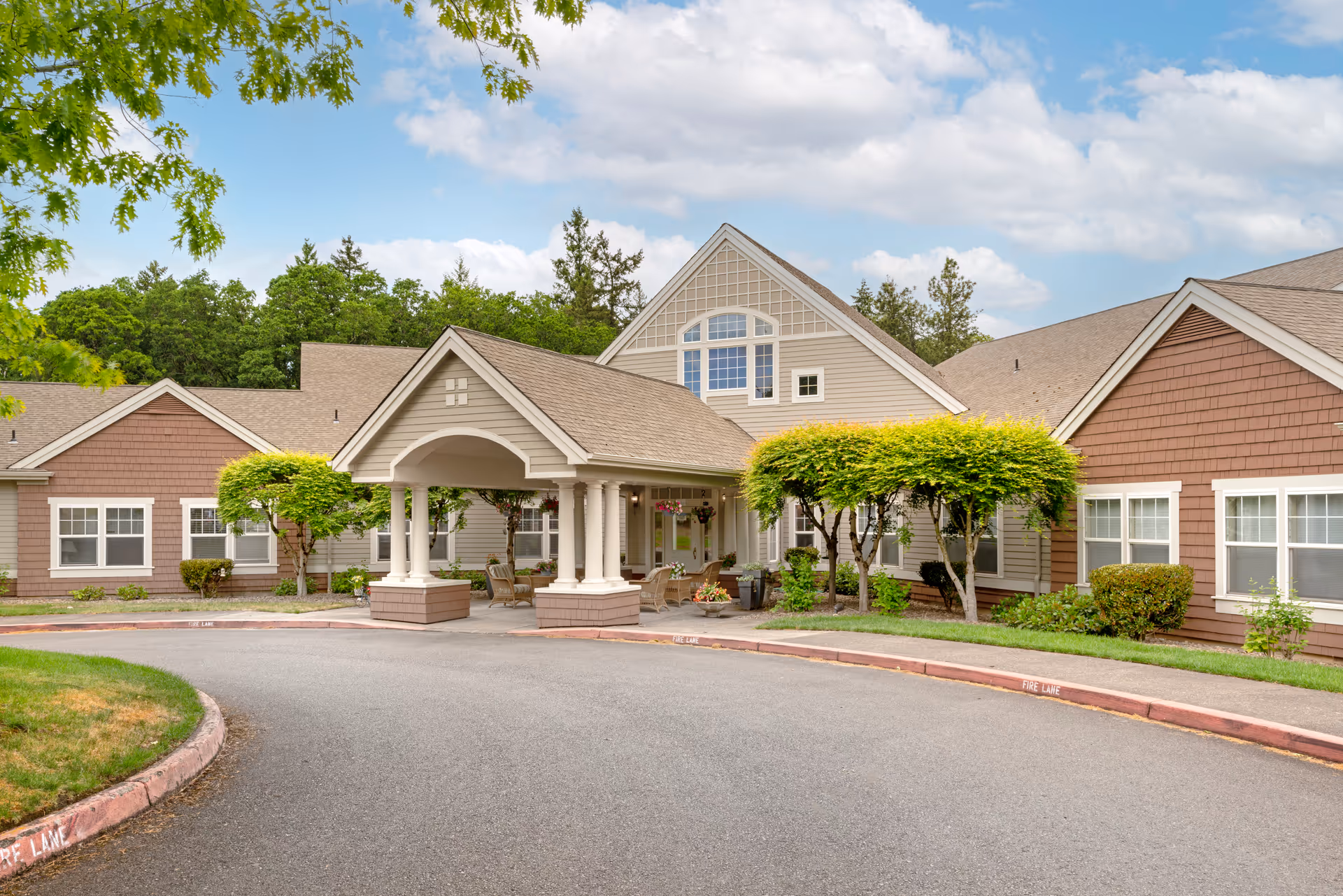 Front entrance of a single-story senior living building with a covered porte-cochere, landscaped trees, and a circular driveway.