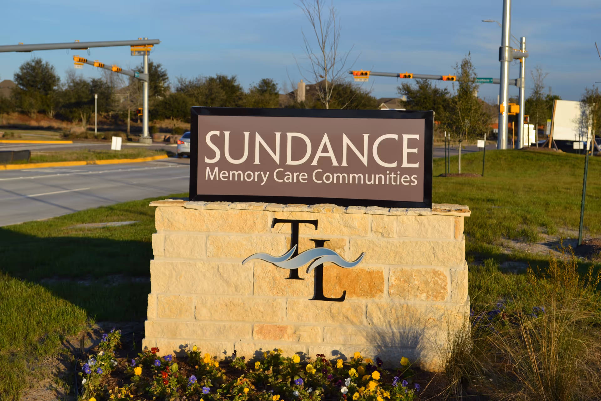 A stone sign at the edge of a grassy area with flowers in front, displaying the text 'SUNDANCE Memory Care Communities' and the letters 'TL' with a wave design, located near a road with traffic lights and trees in the background.