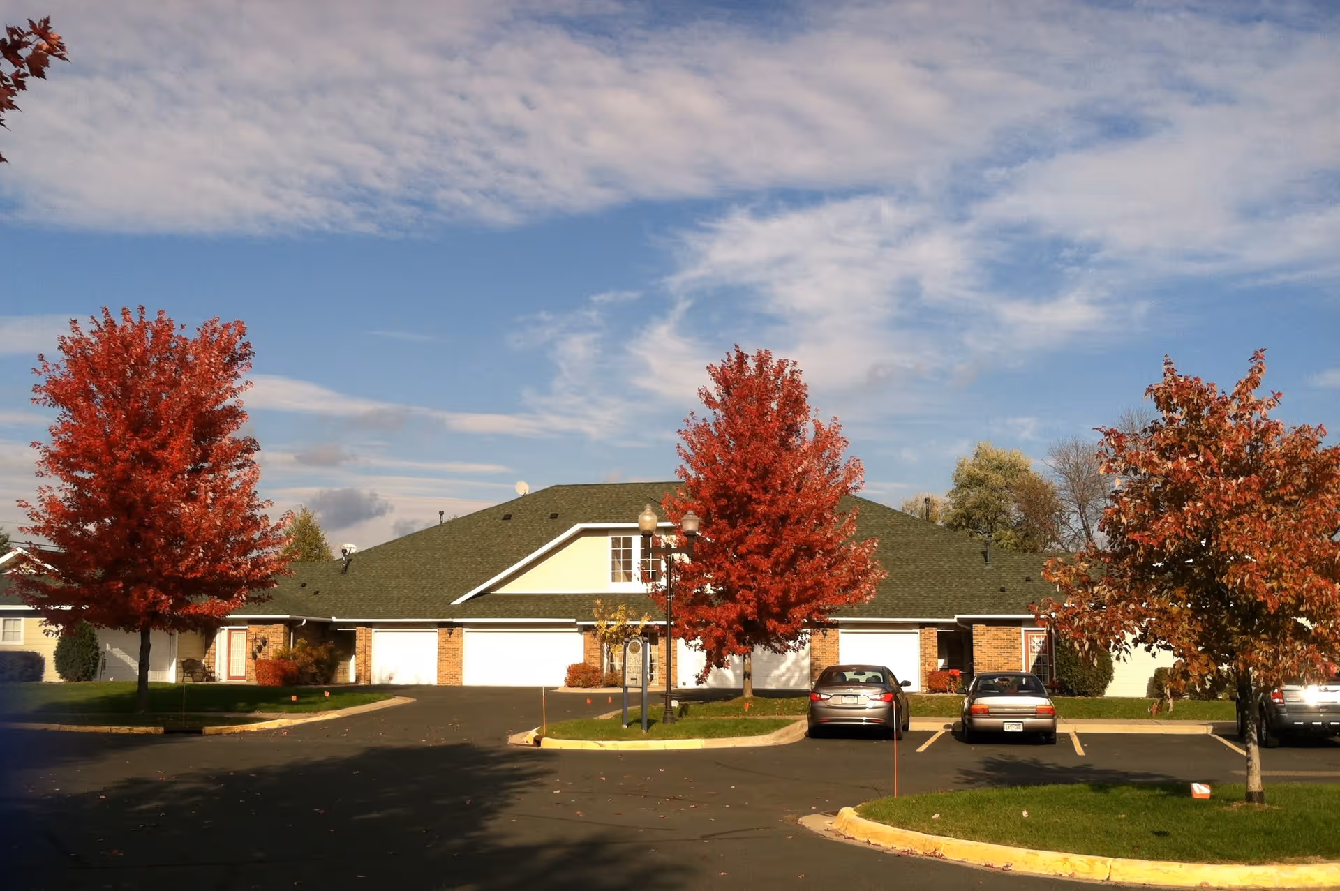 Exterior view of a residential building with a green roof and brick walls, surrounded by a parking lot with several cars and trees with red autumn foliage under a partly cloudy sky.