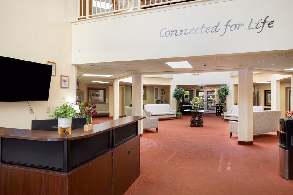 Interior view of a senior living community lobby with a reception desk on the left, a flat screen TV mounted on the wall, and several white cushioned sofas arranged around a central table with a floral arrangement. The carpet is red, and there are plants and framed pictures decorating the space. Above the open area, the phrase 'Connected for Life' is written on the wall.