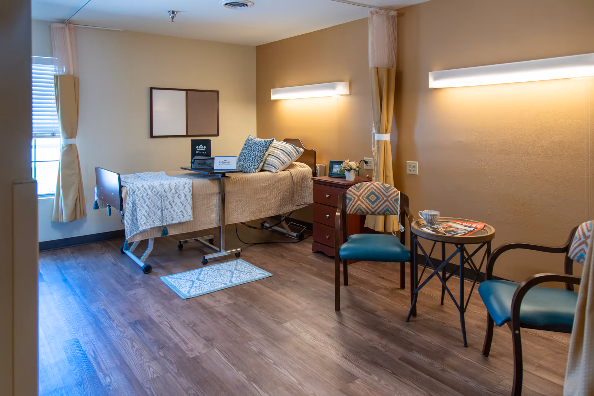 A nursing facility bedroom with a single hospital bed covered with a beige blanket and patterned pillows. Next to the bed is a wooden nightstand with a small flower arrangement and a framed photo. Two chairs with patterned upholstery and blue seats are placed around a small round table holding a magazine and a cup. The room has wood flooring, beige walls, a window with curtains, and soft lighting fixtures above the bed and table.