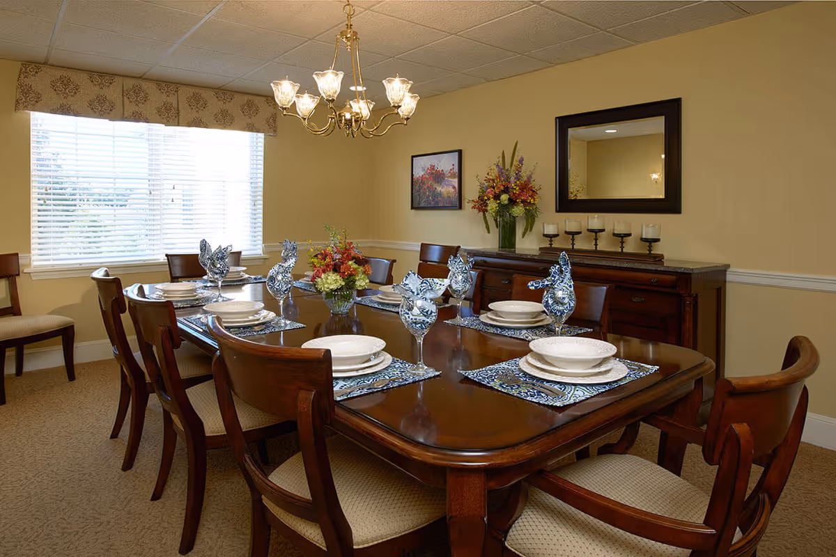 Formal dining room with a polished wooden table set for eight, a chandelier overhead, a window with blinds, and a sideboard with mirror and flowers.