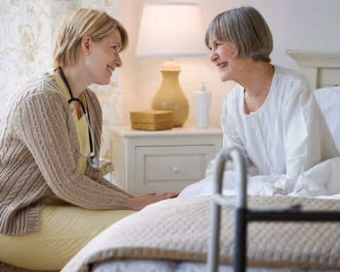A caregiver kneels beside a smiling elderly woman sitting on a bed with a bedside table and lamp in the background.